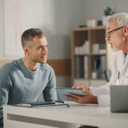 Experienced Middle Aged Family Doctor Showing Analysis Results on Tablet Computer to Male Patient During Consultation in a Health Clinic. Physician Sitting Behind a Desk in Hospital Office.